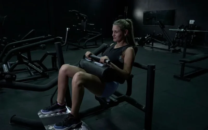 A woman performs leg exercises on a gym machine in a dimly lit environment.