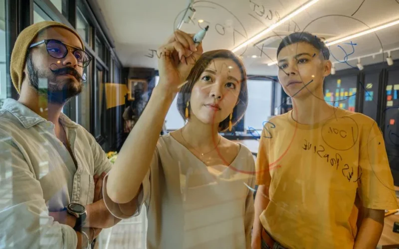 Three colleagues engaged in a strategy meeting, writing ideas on glass in a modern office.