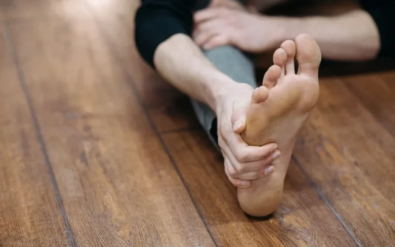 Close-up of a person doing a stretching exercise on a wooden floor, focusing on their bare foot.