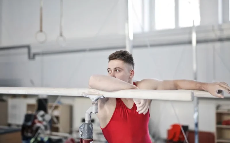 Male gymnast in red attire resting on parallel bars in a gym, showing strength and focus.