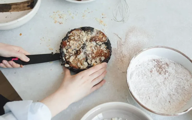 Hands slicing a pear and almond cake in a cozy kitchen setting with flour and whisk.