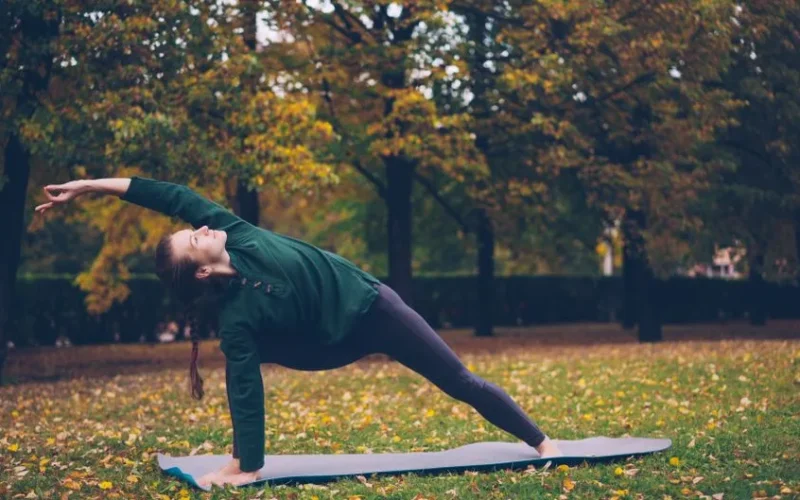 Woman doing a yoga pose on a mat in a park surrounded by autumn trees.