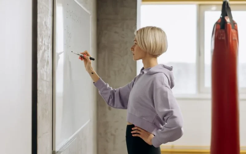 Fit female trainer writes workout plan on a whiteboard in a gym setting. Side view.