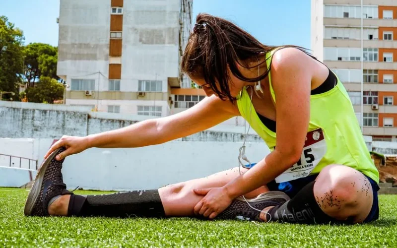 A young woman in a neon vest stretching on grass, preparing for an outdoor workout or running event.
