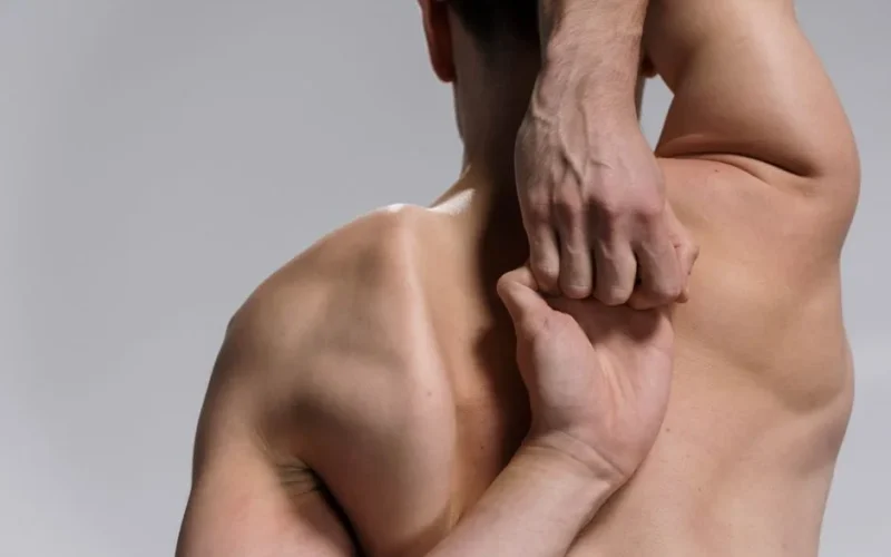 Shirtless muscular man practicing a yoga stretch with hands clasped behind back on a white background.