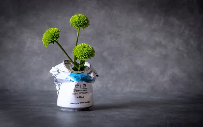 Green pom pom flowers creatively arranged in a crushed vodka can, showcasing innovative recycling.