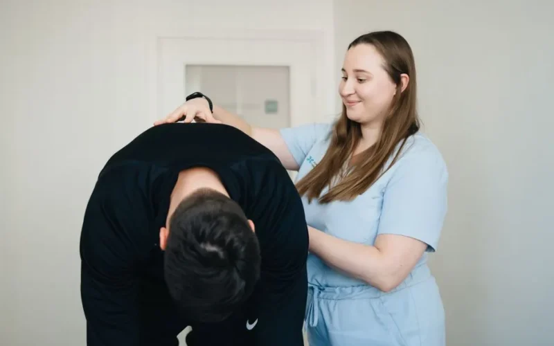 A physiotherapist smiling while assisting a patient with a back exercise in a clinic.