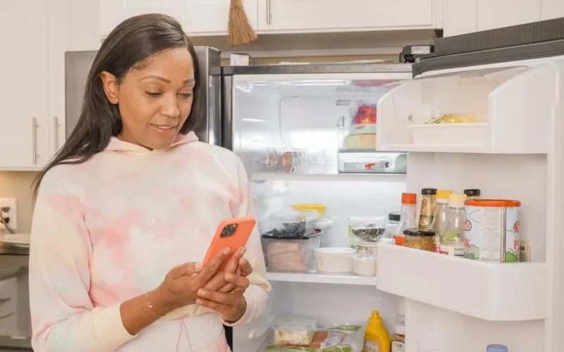 Woman using smartphone for online shopping while standing in the kitchen with an open refrigerator.