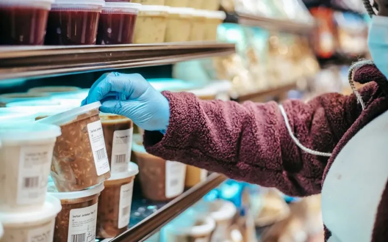 Crop anonymous female customer in protective mask reading label on frozen food in plastic container in grocery store