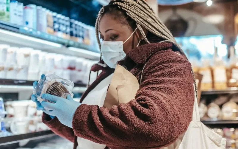 Side view of African American woman in protective gloves and mask reading sticker on product in grocery store