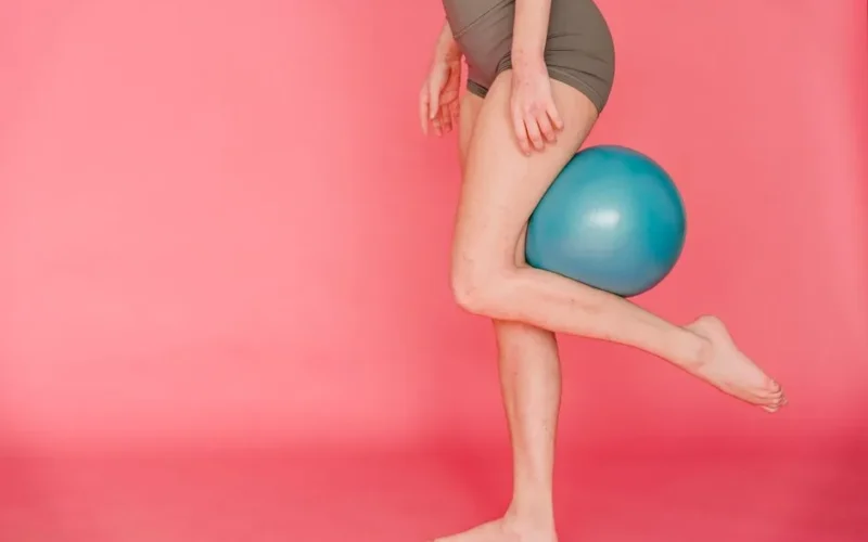 Side view of unrecognizable barefoot slim female gymnast wearing sportive shorts doing exercise with fitness ball while standing on pink background in studio