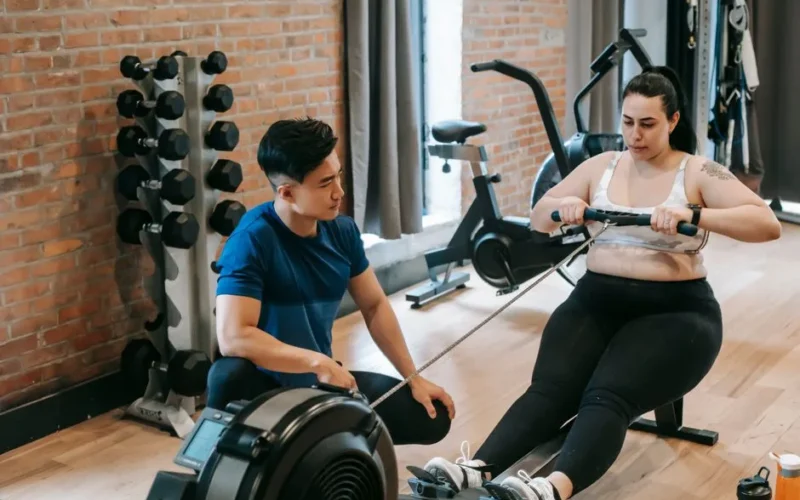 From above of professional ethnic constructor helping young plus size woman working out with rowing machine in sport club