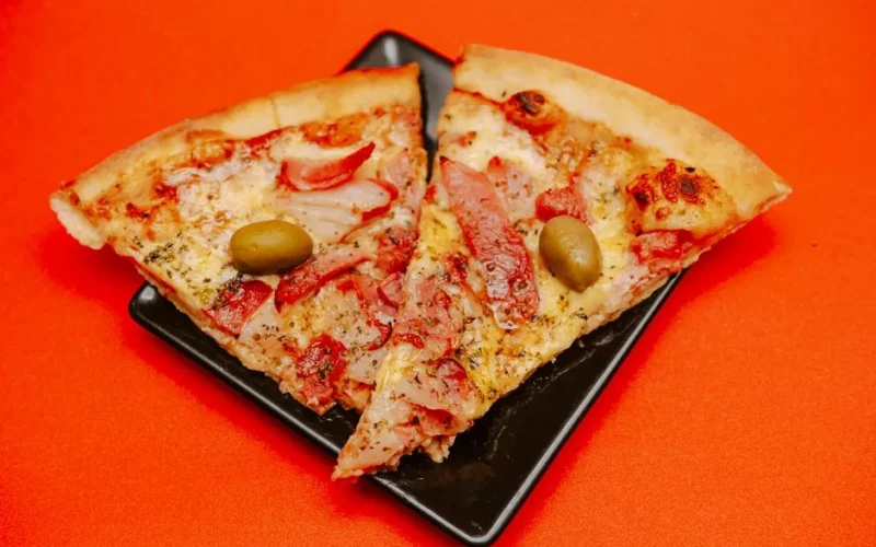 Two savory pizza slices with olives on a black plate against orange backdrop.
