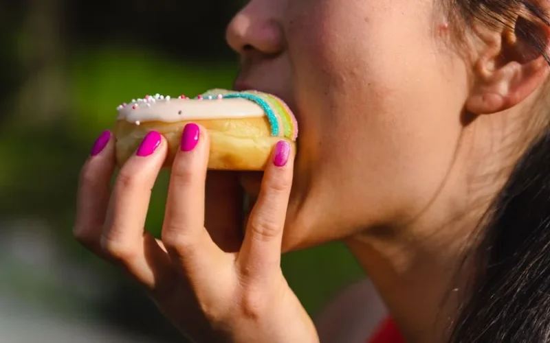 A woman with manicured nails takes a bite of a colorful sprinkled doughnut.