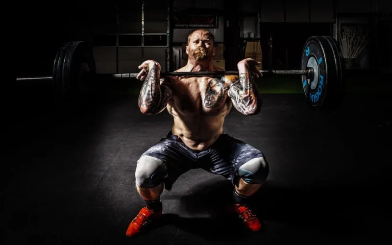 A tattooed man performing a heavy barbell squat in a gym for strength training.