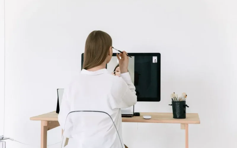 A woman applies makeup at her desk using a computer screen as a mirror.