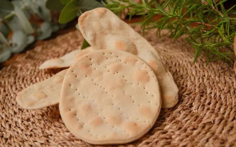 Round and oval flatbreads are placed on a textured woven mat surrounded by fresh green herbs.