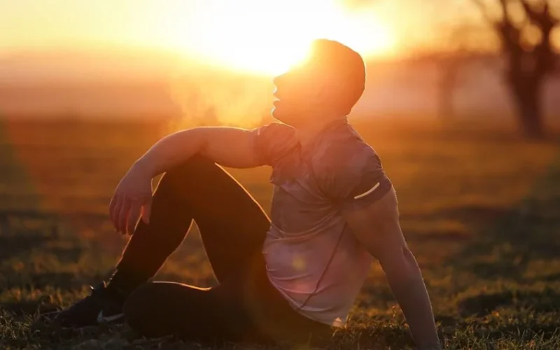 Muscular man sits in rural field at sunset, exhaling visible breath.