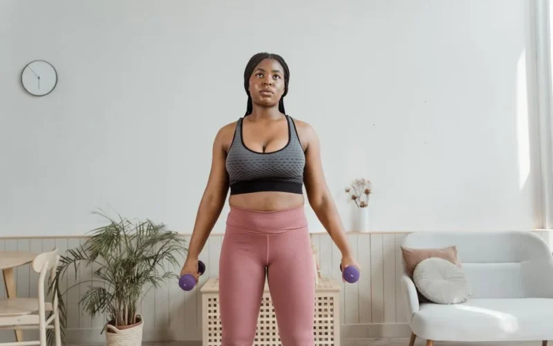 A woman in activewear exercises with dumbbells in a cozy home setting.