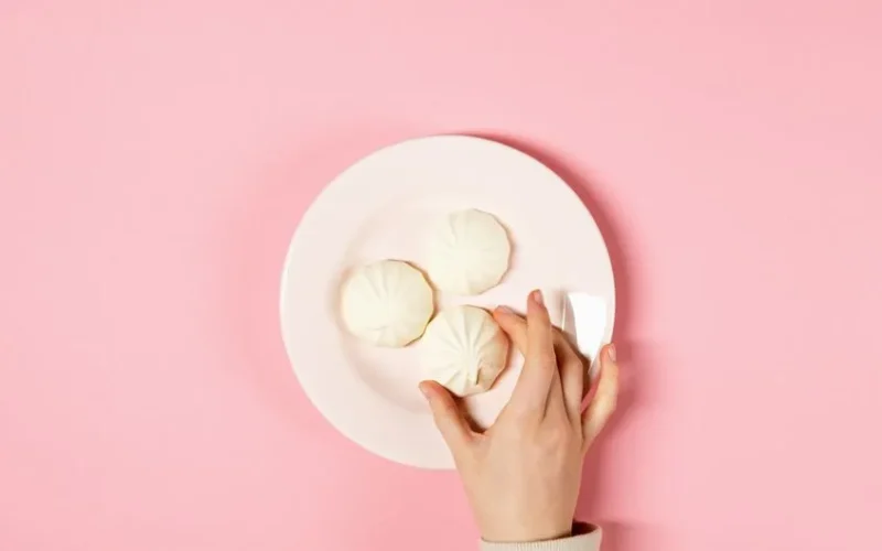 A hand holds a ceramic plate of white meringues against a light pink background.