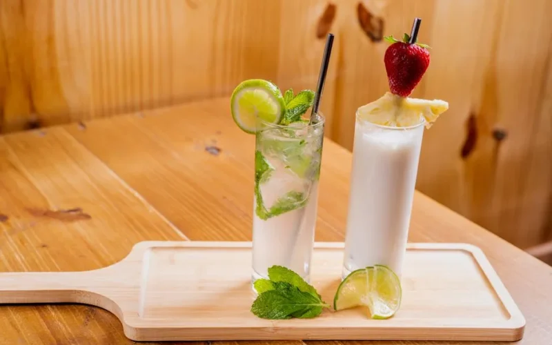 Two tropical cocktails with lime, mint, and fruit garnish on a wooden tray, perfect for summer.
