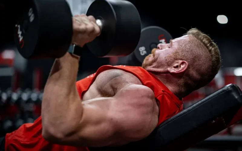 Side view of a muscular man lifting dumbbells on a bench in a gym, focusing on strength and effort.