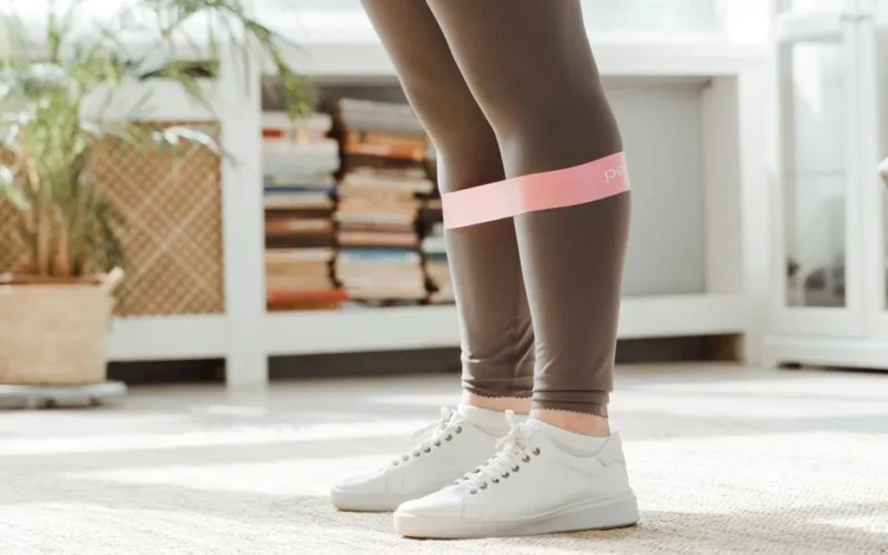Close-up of a person wearing leggings and sneakers, using a resistance band for exercise indoors.