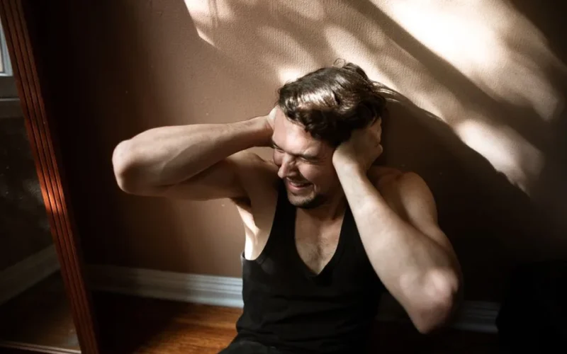 A man sitting on the floor indoors, showing signs of stress by covering his ears.