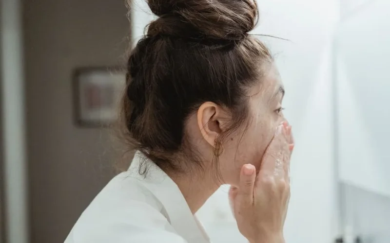 A young woman in a bathrobe follows her skincare routine, washing her face in a bathroom.