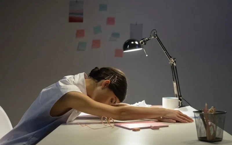 A tired woman rests her head on an office desk under a lamp, symbolizing workplace fatigue.