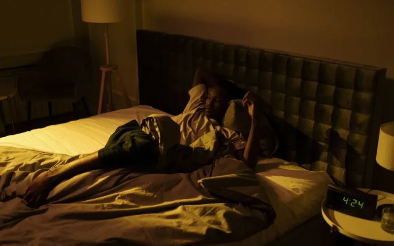 A restless man lying awake on his bed in a dimly lit bedroom at night.