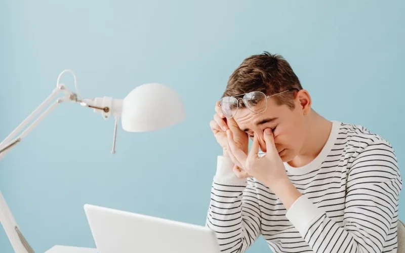 A young man rubs his eyes while working on a laptop, looking tired under a bright desk lamp.