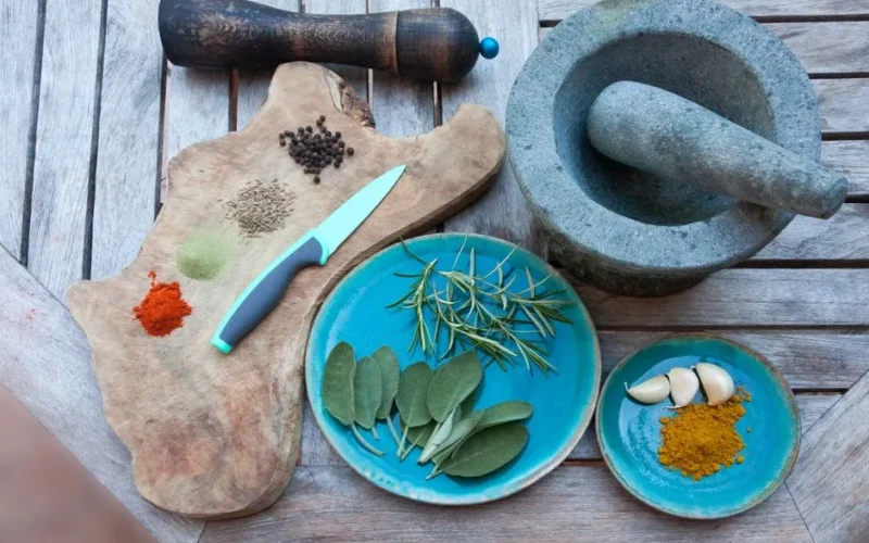 A rustic setup for spice preparation with herbs, spices, garlic, and kitchen tools on wooden table.
