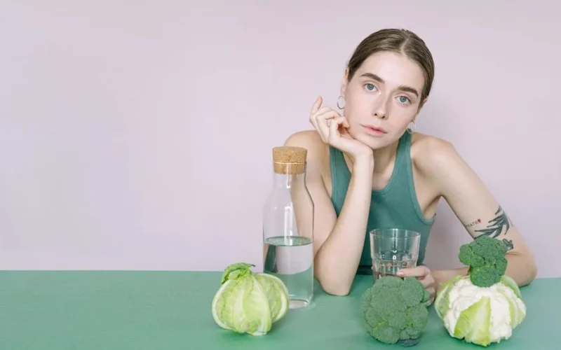 A young woman in a green top sitting with vegetables and a glass of water.
